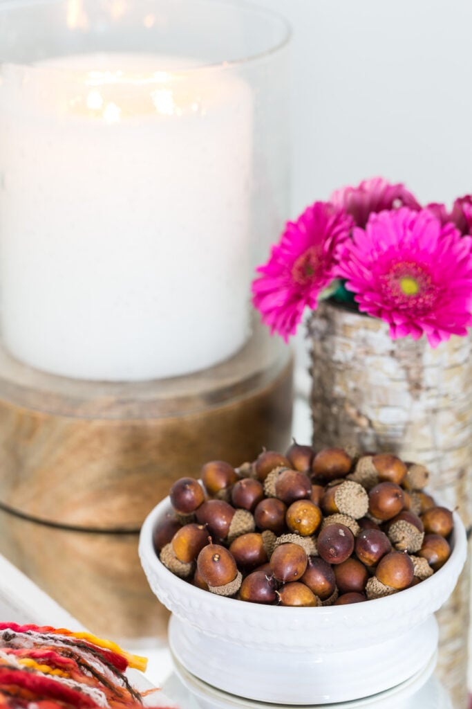 a large candle, bowl of acorns and pink flowers on a side table in a living room decorated for fall