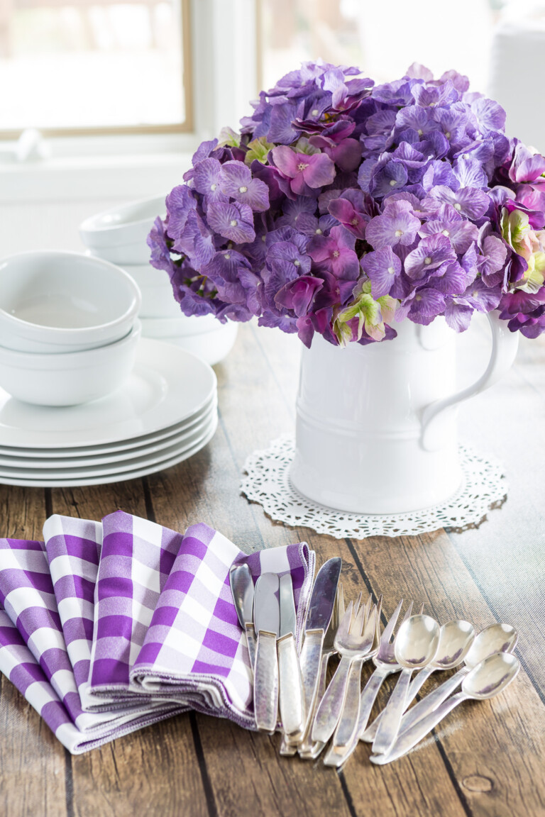 Decorating for spring using White dinnerware and white pitcher filled with purple hydrangeas on a wood table.