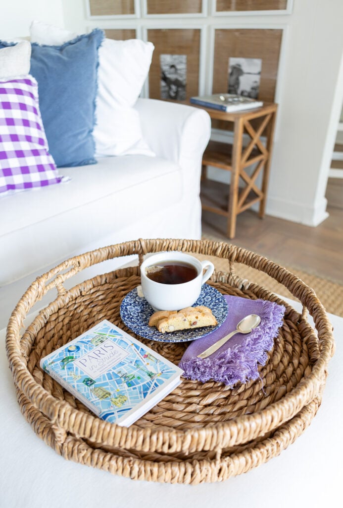 Looking down at a round rattan tray holding a cup of tea, napkin, spoon, a biscotti and a book.