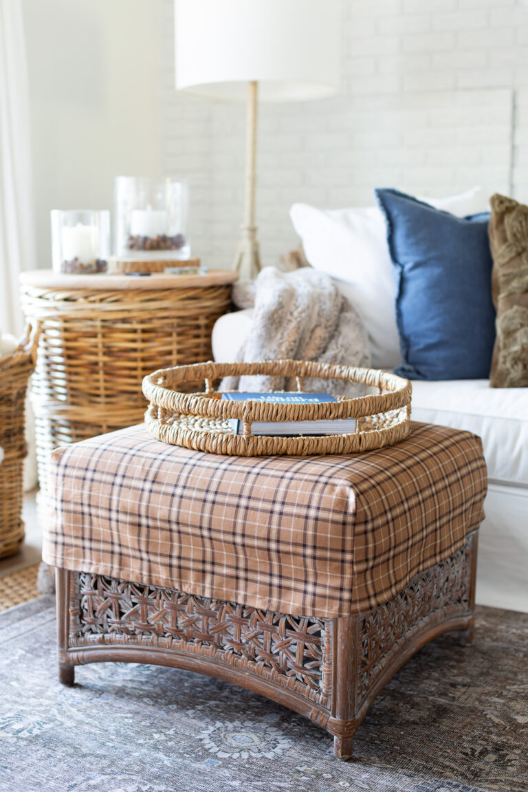 a square rattan ottoman with a brown plaid slipcover on it in from of a white sofa with layers of fall decor on it.