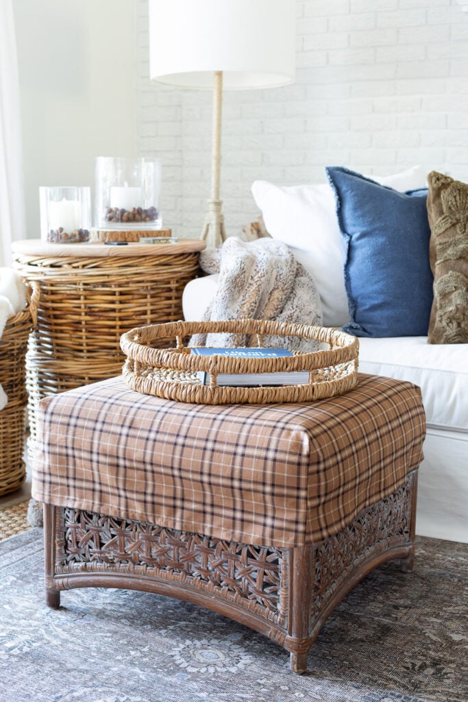 a square rattan ottoman with a brown plaid slipcover on it in from of a white sofa with layers of fall decor on it.