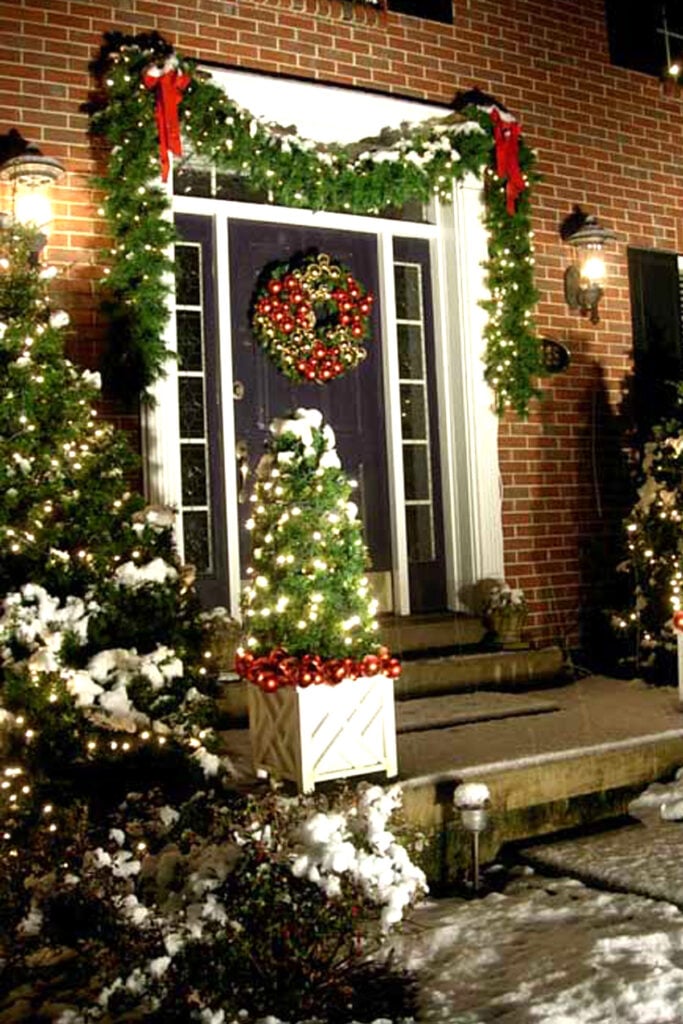 Front exterior of a brick home decorated for Christmas where lit garlands are hug and shrubs decorated with white lights.