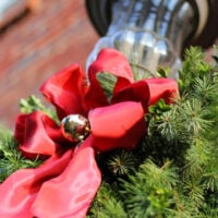 Outdoor porch light decorated for Christmas with greenery and a red ribbon with a gold ornament tied onto it.