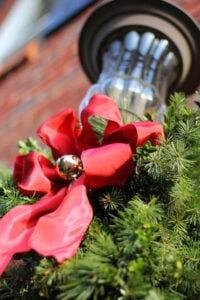 Outdoor porch light decorated for Christmas with greenery and a red ribbon with a gold ornament tied onto it.