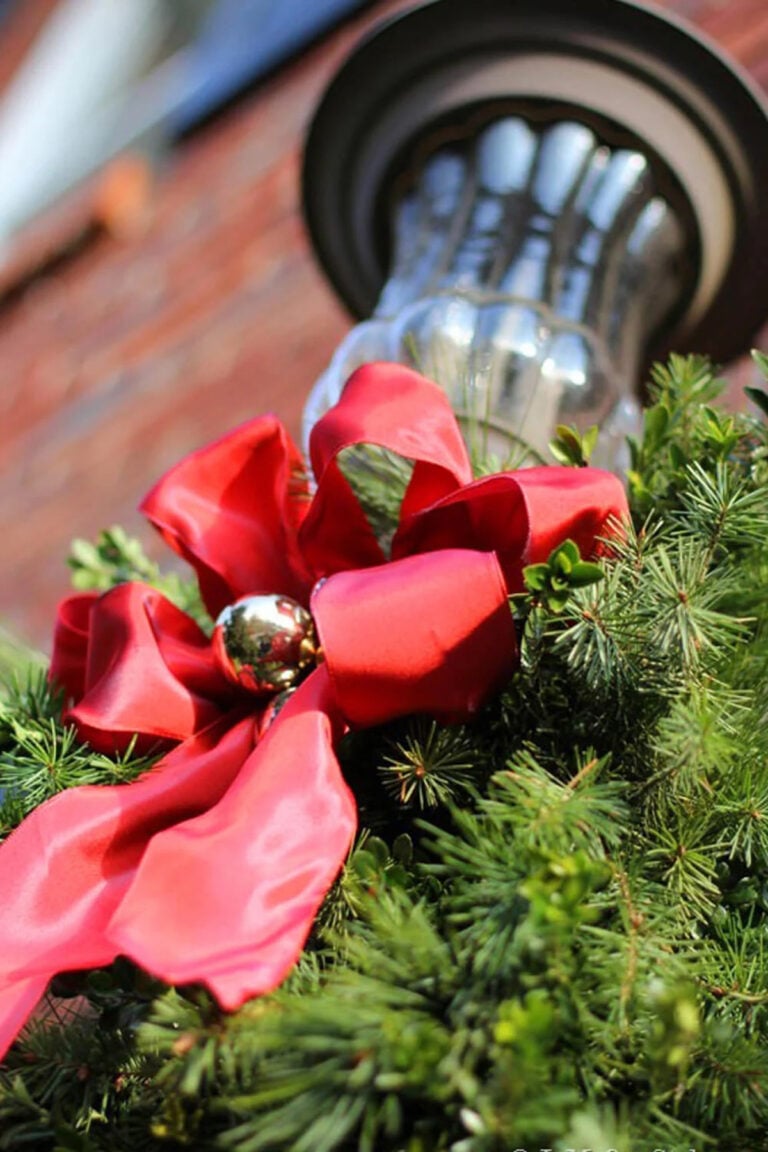 Outdoor porch light decorated for Christmas with greenery and a red ribbon with a gold ornament tied onto it.