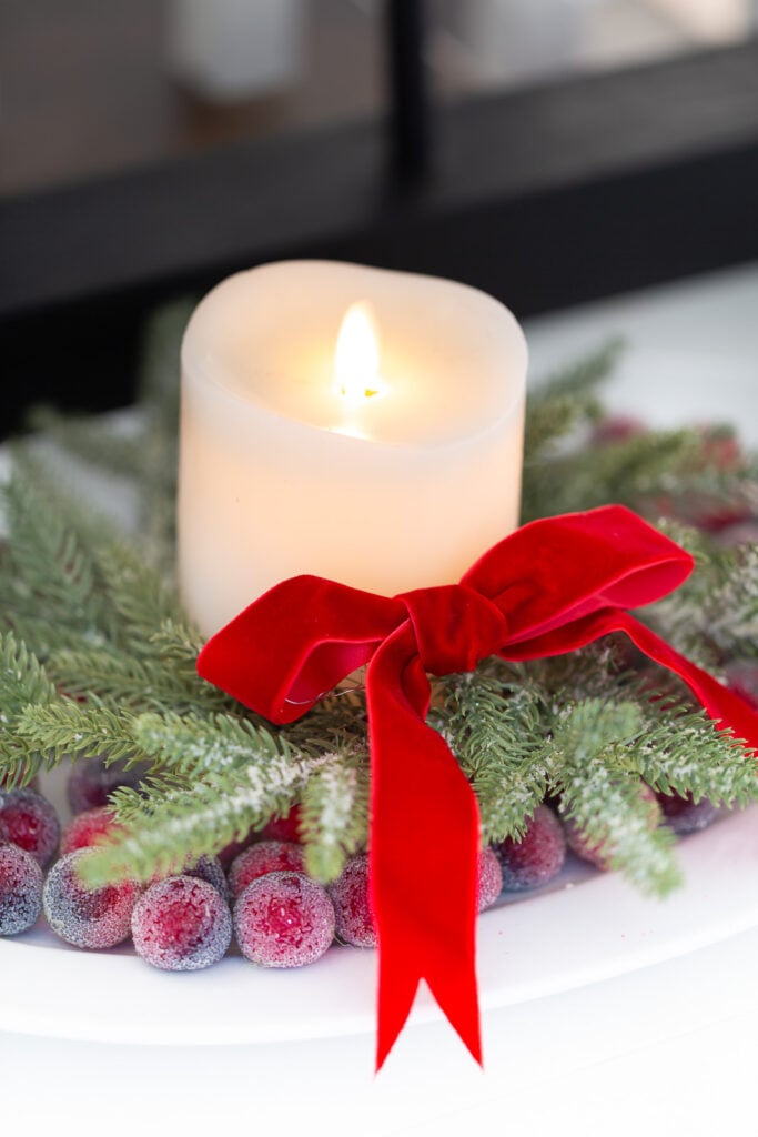 close up of a white candle on a platter with a Christmas wreath around it and sugared cranberries.