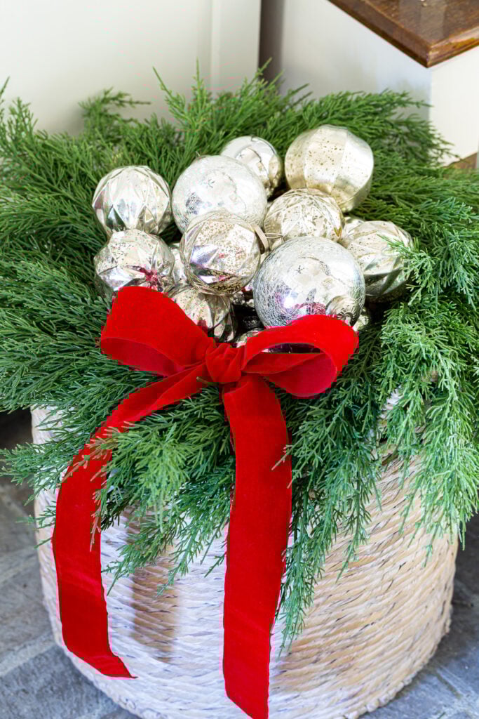 close up of woven floor basket with nestled silver ornaments in a wreath on top with a red velvet bow.