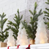 a collection of Glass and glittered Christmas trees with battery-operated candle inside on mantel along with faux pine small trees as Christmas mantel decor.