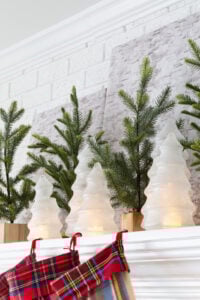 a collection of Glass and glittered Christmas trees with battery-operated candle inside on mantel along with faux pine small trees as Christmas mantel decor.