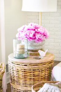Corner of living room with a rounf wicker basket table with a faux concrete planter filled with faux peonies for early spring decor.